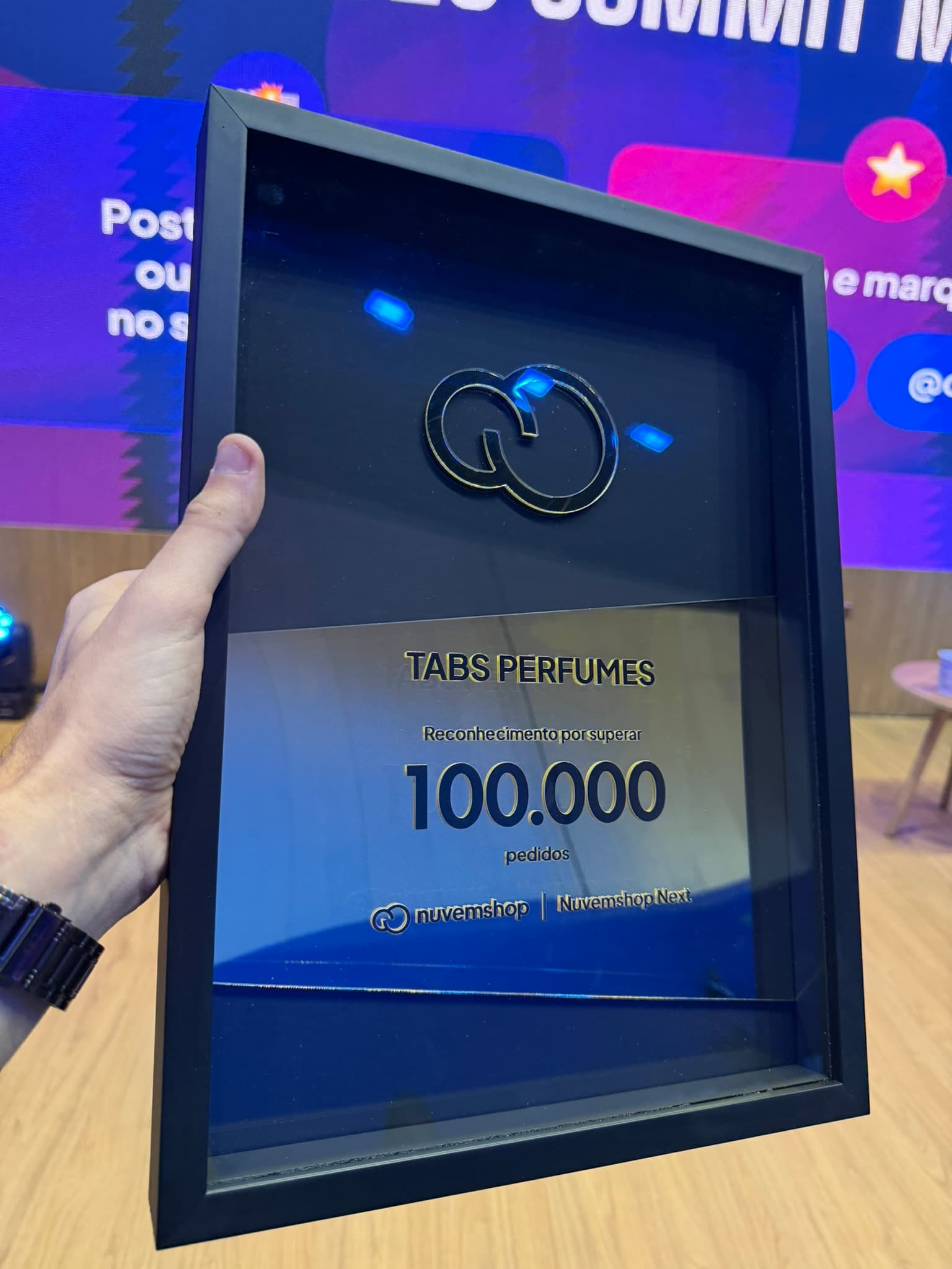Detalhe da placa da Nuvemshop com o nome Tabs Perfumes e o marco de 100.000 pedidos